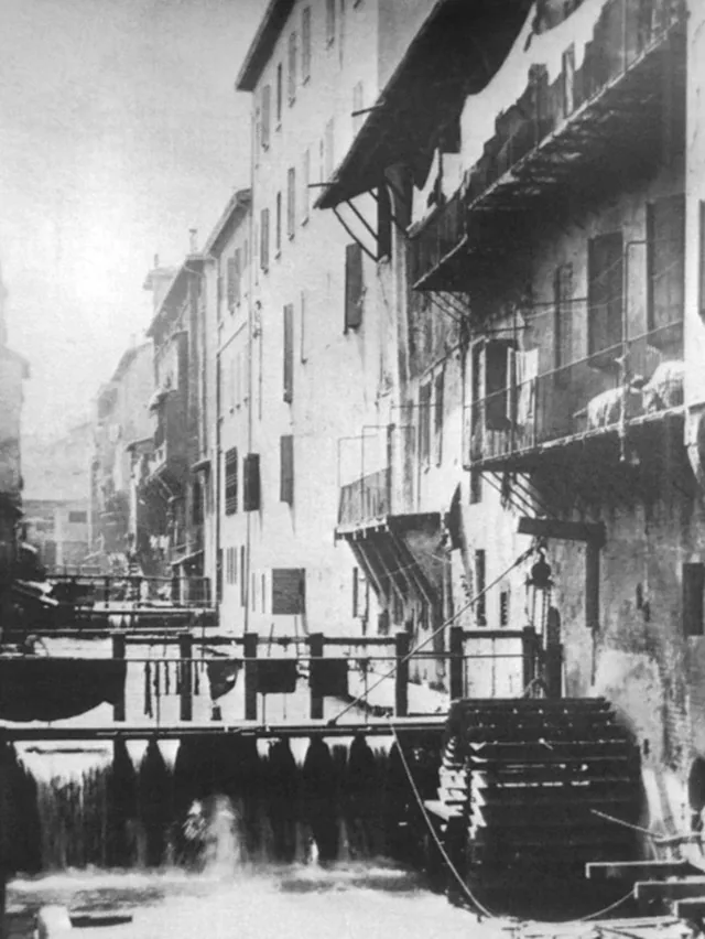 Fotografia storica in bianco e nero del Canale delle Moline a Bologna scoperto, con le case medievali affacciate sull'acqua e una ruota idraulica di mulino in primo piano, prima della copertura novecentesca